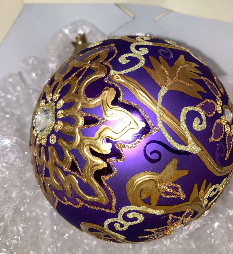 May include: Purple Christmas ornament with gold and glitter accents. The ornament features a floral design.
