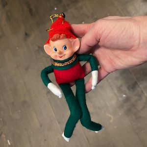 Puede incluir: Un adorno de Navidad de fieltro verde y rojo en forma de elfo con ribete dorado y una cara sonriente. El elfo lleva guantes blancos y un gorro rojo.