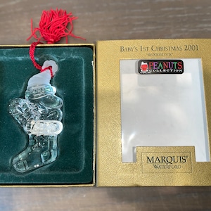 May include: A clear glass Christmas ornament depicting Woodstock from the Peanuts cartoon wearing a Santa hat. The ornament is in a green velvet box with a gold trim. The box has a label that reads "Baby's 1st Christmas 2001" and "Peanuts Collection" with a small image of Woodstock. The box also has a label that reads "Marquis' Waterford".