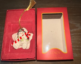 May include: A white ceramic snowman ornament holding a candy cane. The ornament is shaped like a star and has a red and green hat. The ornament is packaged in a red box with the Lenox logo and the words "American by Design".