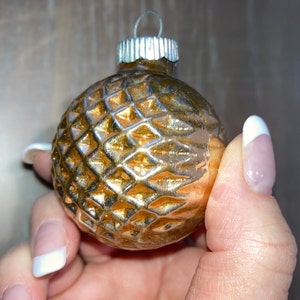 May include: A gold glass Christmas ornament with a textured diamond pattern. The ornament is being held by a hand with white nail polish.