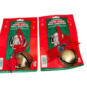 Op de afbeelding: Twee kerstdeurbelgreeters. Elke bel is goudkleurig en bevestigd aan een rood lint en strik. De verpakking is rood en groen met de tekst "Christmas Door Knob Bell Greeter".