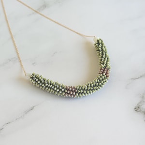 May include: A delicate necklace featuring a curved, textured pendant in shades of green and brown. The pendant is suspended from a thin, gold-colored chain. The necklace is set against a white marble background.