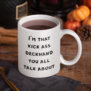 May include: White ceramic coffee mug with black text that reads "I'm that kick ass deckhand you all talk about". The mug is filled with a dark liquid and is sitting on a wooden table with a gray napkin and a gold spoon.