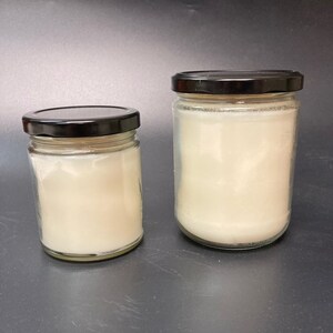 Puede incluir: Dos tarros de vidrio para velas con tapas negras. El tarro m&aacute;s peque&ntilde;o est&aacute; a la izquierda y el m&aacute;s grande a la derecha. Ambos tarros est&aacute;n llenos de cera de vela blanca.