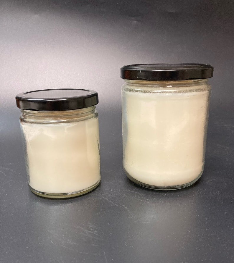 Puede incluir: Dos tarros de vidrio para velas con tapas negras. El tarro m&aacute;s peque&ntilde;o est&aacute; a la izquierda y el m&aacute;s grande a la derecha. Ambos tarros est&aacute;n llenos de cera de vela blanca.