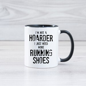 May include: White ceramic coffee mug with a black rim and handle. The mug has black text that reads "I'm not a hoarder I just need more running shoes".
