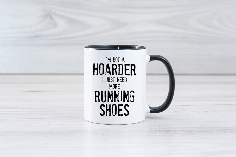 May include: White ceramic coffee mug with a black rim and handle. The mug has black text that reads "I'm not a hoarder I just need more running shoes".