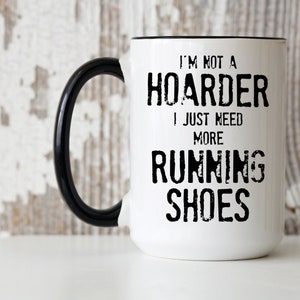 May include: White ceramic coffee mug with a black handle. The mug has black text that reads "I'm not a hoarder I just need more running shoes".