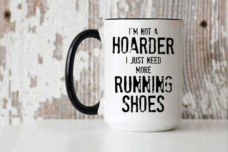 May include: White ceramic coffee mug with a black handle. The mug has black text that reads "I'm not a hoarder I just need more running shoes".