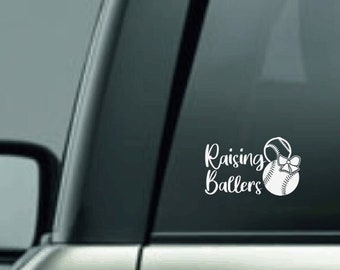 May include: White vinyl decal featuring the text "Raising Ballers" in a script font, accompanied by a baseball with a bow. The decal is designed to be applied to a car window or similar surface.