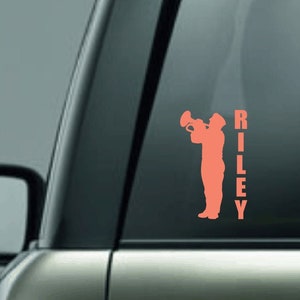 May include: A pink silhouette of a person playing a trumpet with the name "RILEY" written vertically next to them. The image is on a car window.