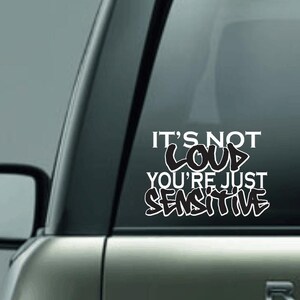 May include: White vinyl decal with the text "IT'S NOT LOUD YOU'RE JUST SENSITIVE" in a graffiti-style font. The decal is applied to a car window, with the vehicle's body visible in the background.