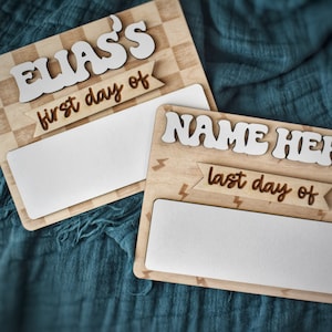 May include: Two wooden signs with blank spaces for writing. The top sign has a checkered background and says "ELIAS'S first day of". The bottom sign has a lightning bolt pattern and says "NAME HERE last day of".