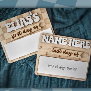 May include: Two wooden signs with a checkered pattern and lightning bolt accents. The top sign says "ELIAS'S first day of" and the bottom sign says "NAME HERE last day of" with the text "this is dry-erase!" below.