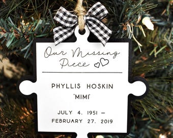 May include: A white and black puzzle-shaped ornament with the text "Our Missing Piece" and two small hearts. The ornament also includes the name "Phyllis Hoskin 'Mimi'" and the dates "July 4, 1951 - February 27, 2019".