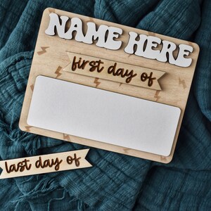 May include: Wooden sign with the text "NAME HERE" in white letters. The sign has a white space for writing in the middle and two banners with the text "first day of" and "last day of" in brown letters.