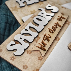 May include: A wooden sign with the name "Savior's" in white letters and the text "first day of" on a banner. The sign has a light brown wood background with a floral pattern.