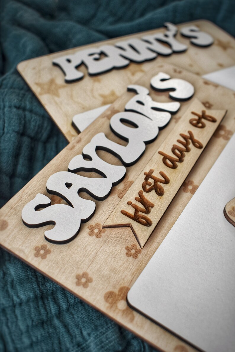 May include: A wooden sign with the name "Savior's" in white letters and the text "first day of" on a banner. The sign has a light brown wood background with a floral pattern.