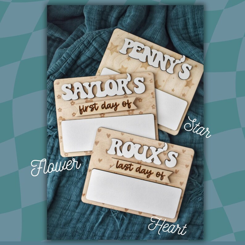 May include: Three wooden signs with white lettering and decorative patterns. The signs say "Penny's", "Sailor's first day of", and "Roux's last day of". The signs are decorated with stars, flowers, and hearts.