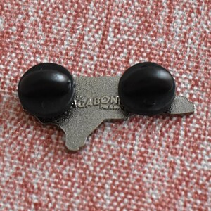 Op de afbeelding: Een zilverkleurige pin in de vorm van een staatsomtrek, met twee zwarte pinruggen. De tekst "GABON" en "PIN SUP" is zichtbaar op de pin. De pin is geplaatst tegen een rood-wit patroon.