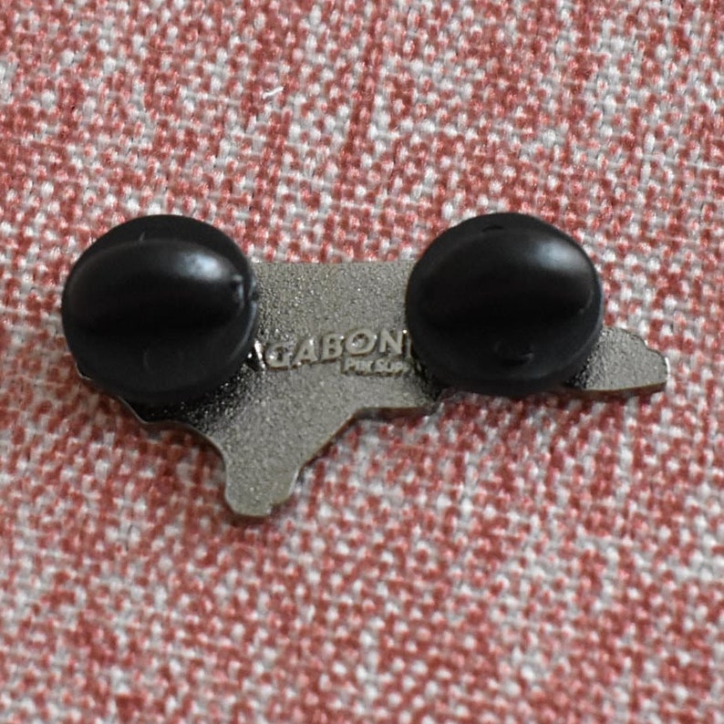 Op de afbeelding: Een zilverkleurige pin in de vorm van een staatsomtrek, met twee zwarte pinruggen. De tekst "GABON" en "PIN SUP" is zichtbaar op de pin. De pin is geplaatst tegen een rood-wit patroon.