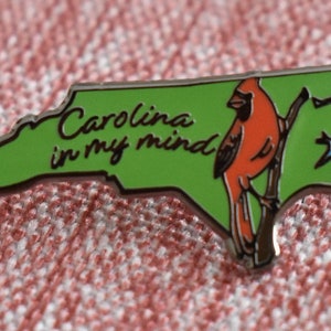 Op de afbeelding: Rechthoekige pin in de vorm van North Carolina, met een groene achtergrond en een rode kardinaal op een tak. De tekst "Carolina in my mind" is in sierlijke letters geschreven.