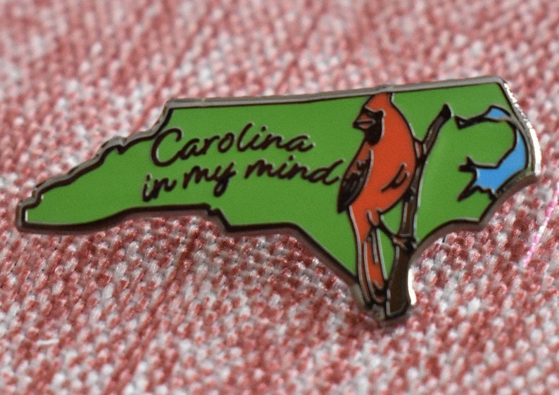 Op de afbeelding: Rechthoekige pin in de vorm van North Carolina, met een groene achtergrond en een rode kardinaal op een tak. De tekst "Carolina in my mind" is in sierlijke letters geschreven.