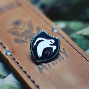 May include: A black and white enamel pin featuring a ghost design with red accents. The pin is set against a brown leather background with the word "ARMY" embossed. The pin has a green border.