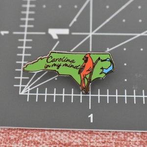 Op de afbeelding: Groene en gouden emaille pin in de vorm van North Carolina, met een rode kardinaal en de tekst "Carolina in my mind". De pin ligt op een grijze ondergrond met meetmarkeringen.