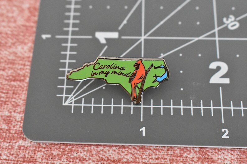 Op de afbeelding: Groene en gouden emaille pin in de vorm van North Carolina, met een rode kardinaal en de tekst "Carolina in my mind". De pin ligt op een grijze ondergrond met meetmarkeringen.
