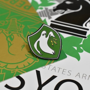 May include: A green and white enamel pin featuring a ghost with lightning bolts on a shield. The pin is set against a background of green and gold designs and text that reads "PER" and "STATES ARMY".