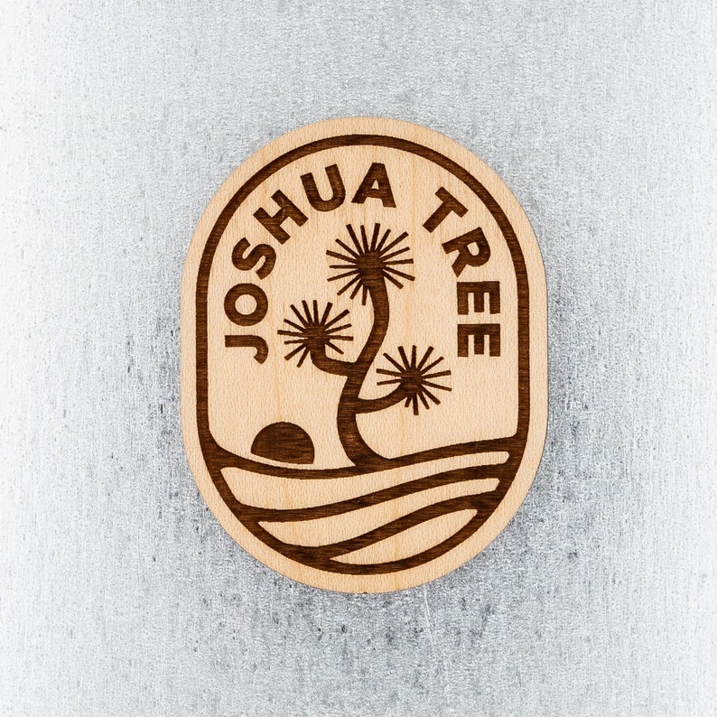 Pu&ograve; includere: Un cartello rotondo in legno con il testo "Joshua Tree" in un cerchio attorno a una silhouette stilizzata di Joshua Tree. L'albero &egrave; marrone e lo sfondo &egrave; in legno chiaro.