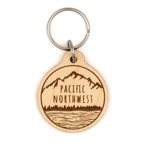 May include: A wooden keychain with a laser-engraved design of mountains, a lake, and trees. The text "PACIFIC NORTHWEST" is engraved on the keychain.
