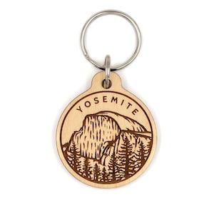 May include: A wooden keychain with a laser-engraved image of Half Dome, a famous rock formation in Yosemite National Park, California. The keychain is circular and has the word "Yosemite" engraved around the edge.