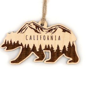 May include: Wooden ornament shaped like a California bear with a mountain range and forest silhouette inside. The ornament is brown and has the word "CALIFORNIA" engraved on it.