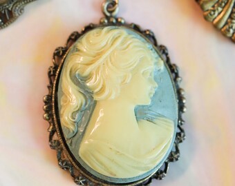 May include: A vintage cameo pendant featuring a woman's profile in white against a blue background. The cameo is set in a gold-toned metal frame with intricate details.