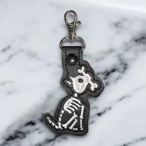May include: Black leather keychain featuring a white embroidered skeleton design. The keychain has a silver-colored clasp and a black snap closure. The skeleton design is detailed and whimsical.