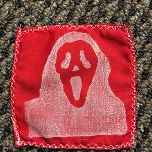 Pu&ograve; includere: Una toppa in tessuto rosso con un disegno di maschera Scream bianca. La toppa quadrata ha un bordo cucito bianco. La maschera Scream, con la bocca aperta, &egrave; centrata.