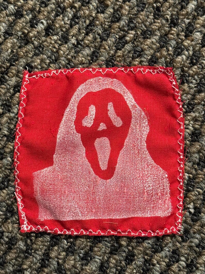 Pu&ograve; includere: Una toppa in tessuto rosso con un disegno di maschera Scream bianca. La toppa quadrata ha un bordo cucito bianco. La maschera Scream, con la bocca aperta, &egrave; centrata.