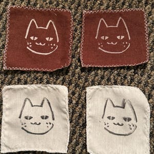 Może przedstawiać: Cztery kwadratowe podkładki z tkaniny. Dwie są w kolorze głębokiego czerwono-brązowego z białym wzorem twarzy kota. Pozostałe dwie są białe z czarnym wzorem twarzy kota. Wszystkie cztery podkładki mają białą, szytą obwódkę.