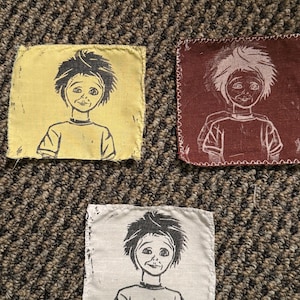 Puede incluir: Tres parches de tela con una ilustración de dibujos animados de un niño con pelo puntiagudo. Los parches son amarillos, granates y blancos, cada uno con un contorno negro o blanco de la imagen. Los parches son cuadrados y tienen un borde cosido.