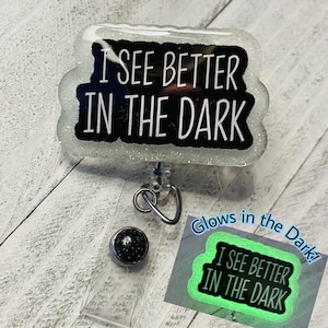 May include: A white glitter badge reel with the text "I SEE BETTER IN THE DARK" in black. The badge reel has a retractable cord and a button with a black and glittery design. A smaller image shows the badge glowing green.
