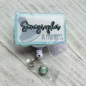 May include: A light blue badge holder with glitter, featuring the text "Sonographer in Progress". The badge holder is attached to a retractable badge reel with a light blue and green glitter charm.