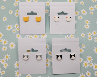 Puede incluir: Cuatro pares de pendientes de botón con forma de gato, presentados en cartulina blanca. Los pendientes son amarillos, blancos, grises y negros.