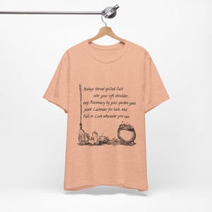 May include: A peach-colored short-sleeve t-shirt featuring a black ink-style illustration of a broom, candles, and a cauldron. The text reads: Always throw spilled Salt over your left shoulder, keep Rosemary by your garden gate, plant Lavender for luck, and Fall in Love wherever you can.