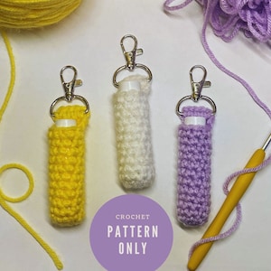 Könnte beinhalten: Drei gehäkelte Lippenbalsam-Halter mit Schlüsselanhängern. Die Halter sind gelb, weiß und lila. Das Bild zeigt einen Häkelhaken und Garn.