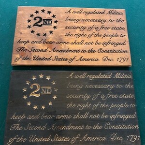 Puede incluir: Dos letreros de madera con la Segunda Enmienda de la Constituci&oacute;n de los Estados Unidos grabada en ellos. El texto es negro y el fondo es de madera. El letrero superior es de madera marr&oacute;n claro y el letrero inferior es de madera marr&oacute;n oscuro. Ambos letreros tienen un dise&ntilde;o de estrellas doradas alrededor del n&uacute;mero "2ND" en la esquina superior izquierda.