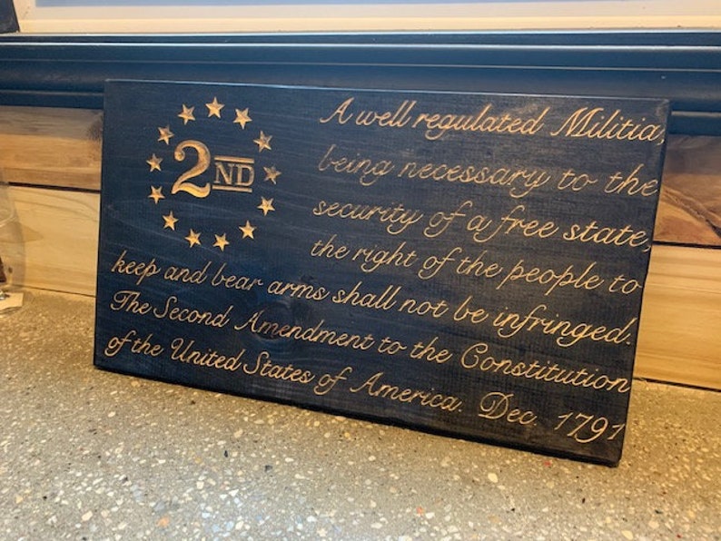 Puede incluir: Un letrero de madera oscura con letras doradas que dice "A well regulated Militia, being necessary to the security of a free state, the right of the people to keep and bear arms shall not be infringed. The Second Amendment to the Constitution of the United States of America. Dec. 1797". El letrero tambi&eacute;n tiene un dise&ntilde;o de estrellas doradas con el n&uacute;mero "2ND" en el centro.
