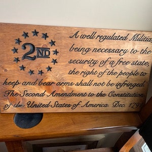 Puede incluir: Un letrero de madera con el texto "A well regulated Militia, being necessary to the security of a free state, the right of the people to keep and bear arms shall not be infringed. The Second Amendment to the Constitution of the United States of America. Dec. 1791" y un c&iacute;rculo con estrellas alrededor del n&uacute;mero "2ND" en el centro.